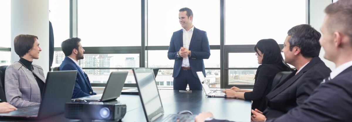 Business professionals in conference room sitting at a table with their computers out. Their attention is to the man at the head of the table speaking